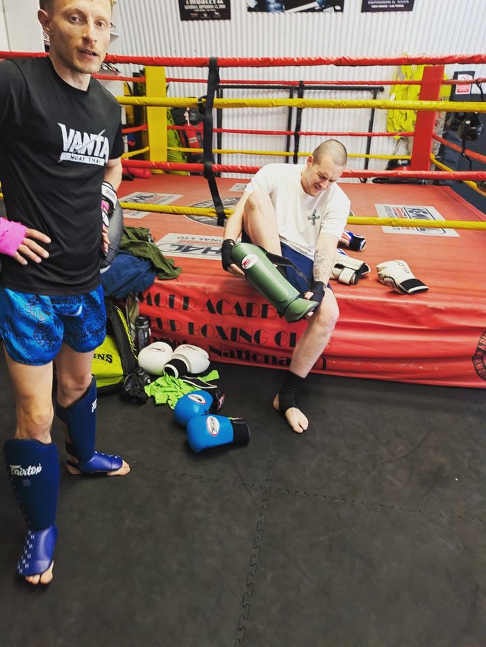Coach in a Vanta T-shirt beside the ring as a student puts on shin guards